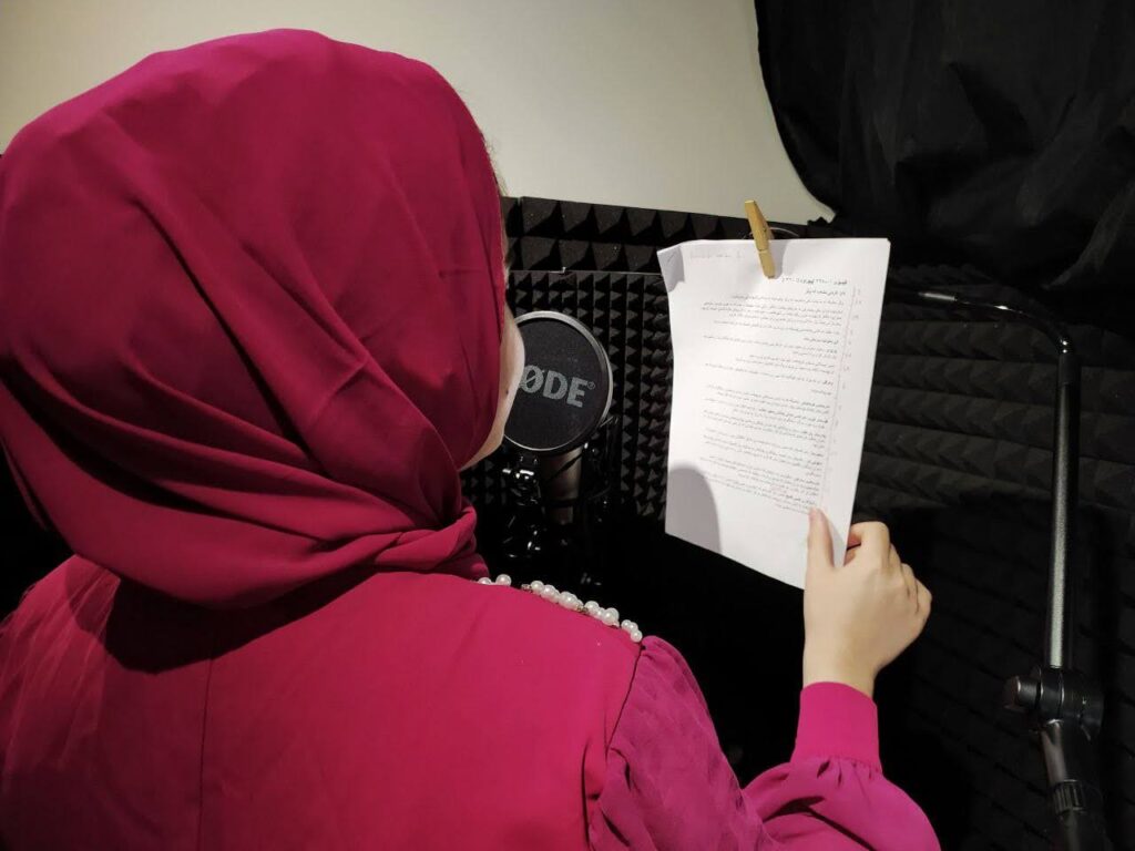 Behind the shoulder shot of a female in a headscarf reading a script in front of a microphone in a soundbooth.